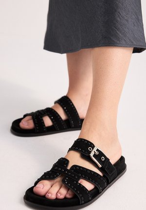 Pies con sandalias de ante negras con tiras tachonadas y hebilla plateada, combinadas con una falda negra sobre un fondo sencillo.