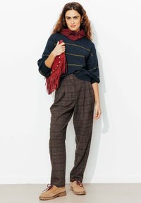 Pull marin à rayures jaunes, pantalon à carreaux marron, et chaussures beiges à lacets. Accessoire : sac rouge à franges avec détails en laiton.