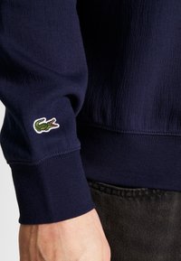 Marinblå stickad sweatshirt med ribbade ärmar, som har en broderad grön krokodil-logotyp på ärmen och en texturerad yta.
