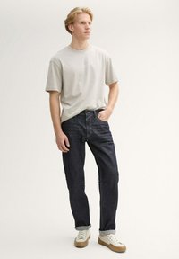 Joven de pie con una mano en el bolsillo, vistiendo una camiseta beige clara, jeans azul oscuro con puños y zapatillas blancas con suelas beige.