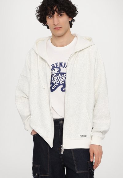 Sweat à zip blanc avec une texture motif, doté d'une capuche à cordon de serrage et de poignets côtelés. Associé à un t-shirt graphique blanc et un pantalon sombre.