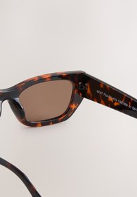Sonnenbrille im Schildpattstil mit braunen Gläsern, rechteckigem Rahmen und schlankem Design. Text am Bügel: "NEXT F24-523-013 FS01056 01 D91."
