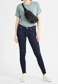 Černá crossbody taška s zipovým uzávěrem, nošená přes světle modrou košili a tmavé slim džíny, doplněná béžovými teniskami.