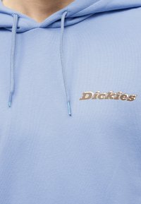 Ανοιχτό μπλε φούτερ με κορδόνια και καφέ λογότυπο «Dickies» στο στήθος, που φοριέται από άτομο που φαίνεται από το λαιμό και κάτω.