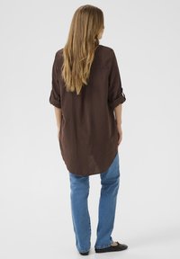 Ženska z dolgimi lasmi, oblečena v rjavo majico z dolgimi rokavi in modre kavbojke, stoji obrnjena stran na belem ozadju.