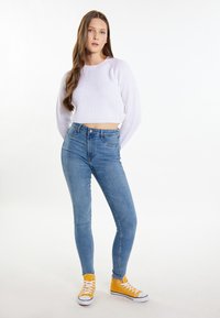 Fehér bordás, cropped pulcsi, magas derekú kék skinny farmer és élénk sárga vászon magasszárú sportcipő fehér fűzővel. Egyszerű dizájn, laza stílus.