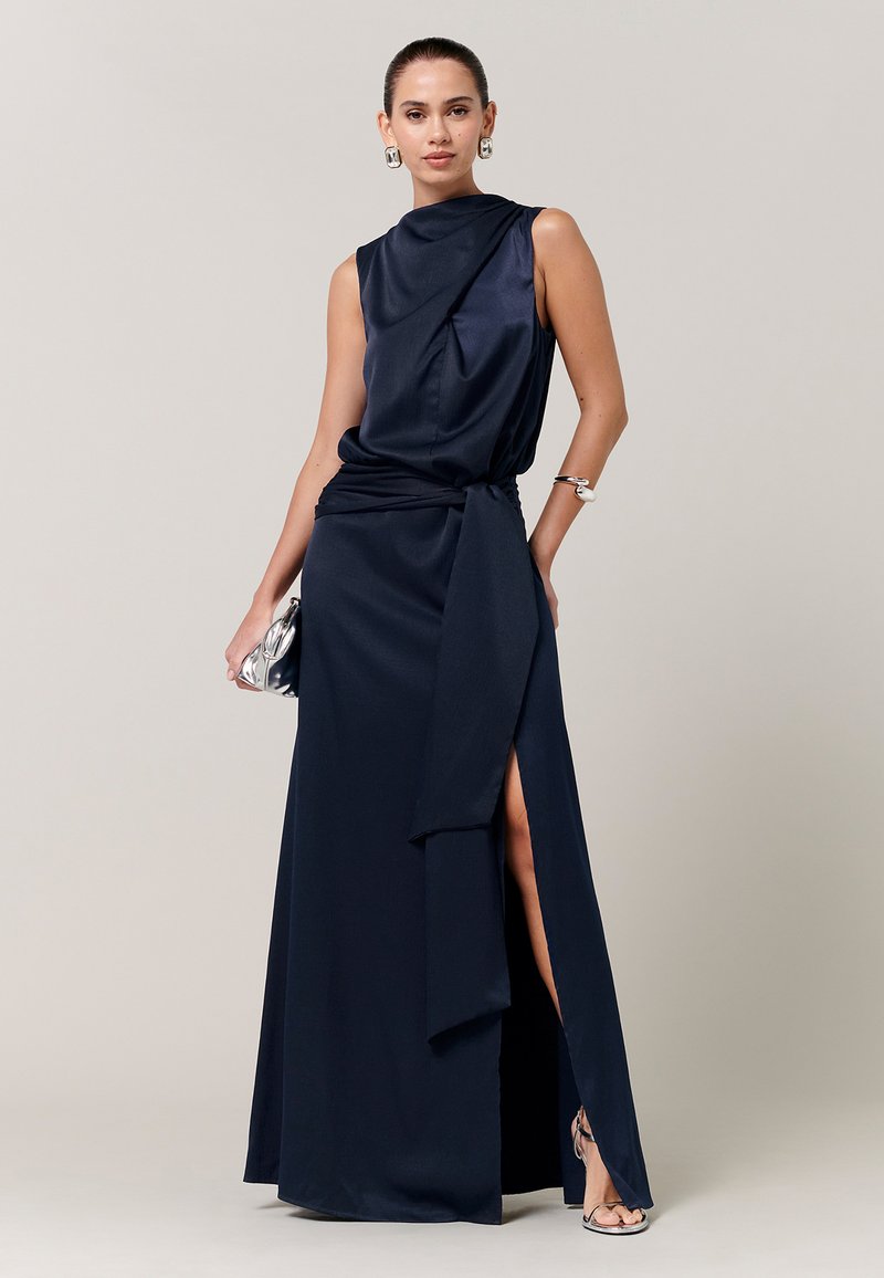 Žena u mornarskoplavoj haljini bez rukava s visokim prorezom, srebrnim potpeticama, velikim srebrnim naušnicama, srebrnom narukvicom i drži malu srebrnu torbicu.