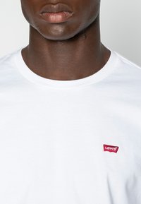 White cotton T-shirt with a round neckline featuring a small red embroidered Levi's logo on the left chest. Smooth texture.