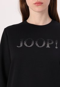 Black sweatshirt featuring a round neckline and "JOOP!" in silver rhinestones across the front. Soft cotton fabric with a smooth texture.