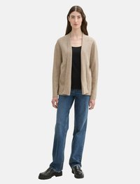 Cardigan beige à manches ouvertes en matériau doux, associé à un haut noir, un jean droit bleu et des chaussures noires imposantes. Design sobre et fonctionnel.