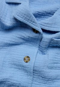 Lichtblauw button-up shirt gemaakt van textuurstof, met goudkleurige knopen en een omgeslagen kraag met zichtbare stiksels.