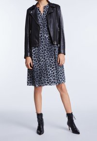 Osoba ubrana w czarną skórzaną kurtkę, niebieską sukienkę w panterkę i czarne sznurowane botki, stojąca na jednolitym tle.