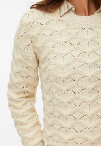 Suéter crema de punto con un patrón de ondas texturizado y puños acanalados. Presenta un escote redondo y hilo entrelazado para ventilación.
