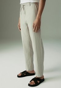 Pantaloni in lino grigio chiaro con vestibilità rilassata, dotati di vita con cordoncino e tasche laterali, abbinati a sandali neri con cinturini incrociati.