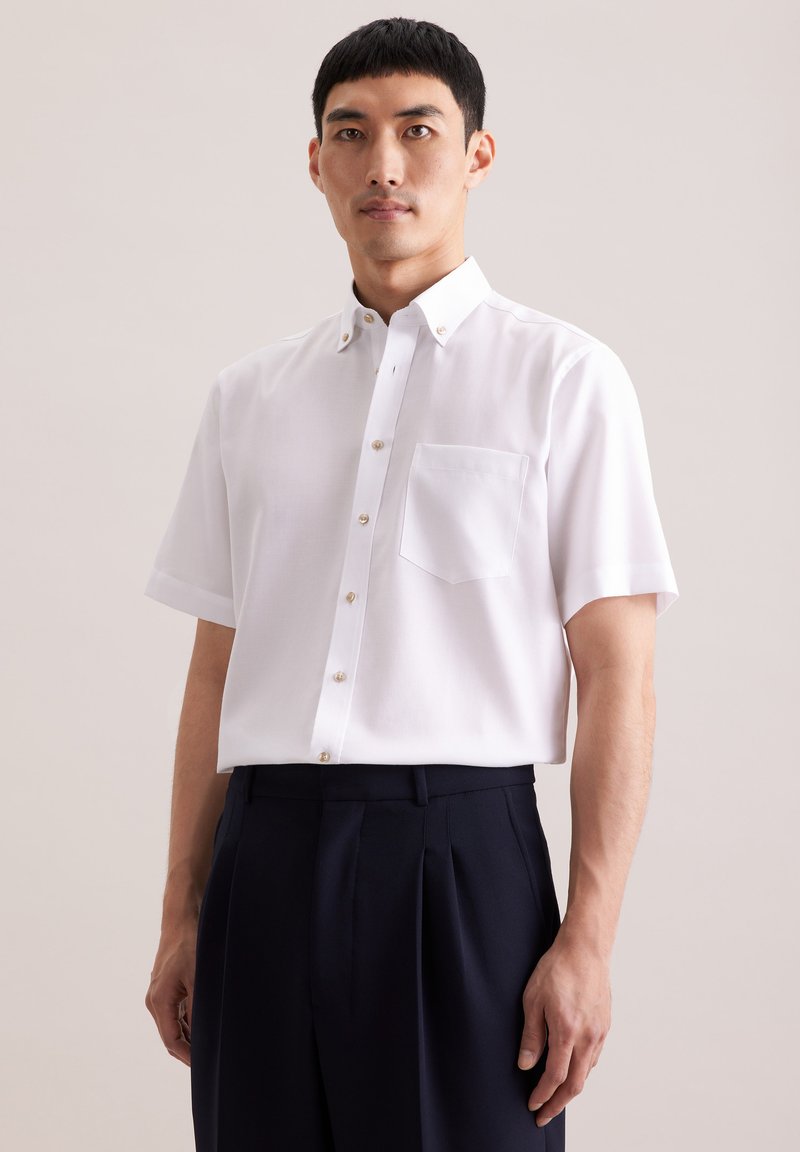 Homme portant une chemise blanche à manches courtes avec poche poitrine et un pantalon noir plissé, sur un fond uni.