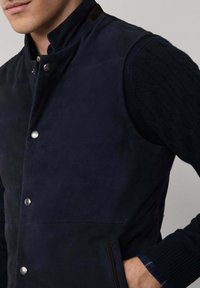 Mornarska jakna s kopčanjem na srebrne dugmadi, s visokim ovratom i kontrastnim smešnim naglaskom na ramenima, uparena s teksturiranim tamnim džemperom.