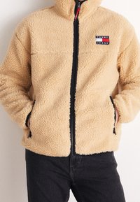 Giacca zip-up beige e pelosa con il logo Tommy Jeans sul petto, indossata con pantaloni neri, mani nelle tasche della giacca.