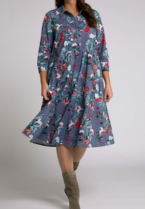 Femme portant une robe midi boutonnée grise à motif floral avec des manches trois-quarts et des bottes mi-mollet en daim taupe, debout devant un fond uni.
