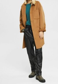 Brauner Wollmantel mit großer Fronttasche, tealfarbiges Rundhalsoberteil, schwarze Kunstlederhose und olivgrüne Stiefeletten. Lässiges Ensemble.