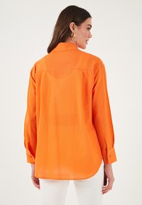 Oranžová košile s dlouhým rukávem, s knoflíkovou manžetou, zadním klopou a uvolněným střihem. Vyrobena z lehkého materiálu s hladkou texturou.