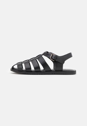 Sandal aperto in pelle nera con diverse cinghie orizzontali e chiusura a fibbia su una suola piatta, su sfondo bianco.