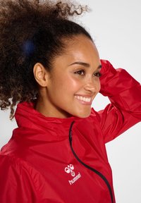 Lächelnde Frau mit lockigem Haar, die eine rote Jacke mit Reißverschluss trägt, richtet ihr Haar mit einer Hand und schaut nach rechts vor einem schlichten Hintergrund.