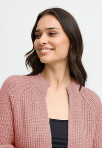 Ljusrosa stickad cardigan med ribbat mönster, med rund krage och öppen design, bärs över en svart topp.