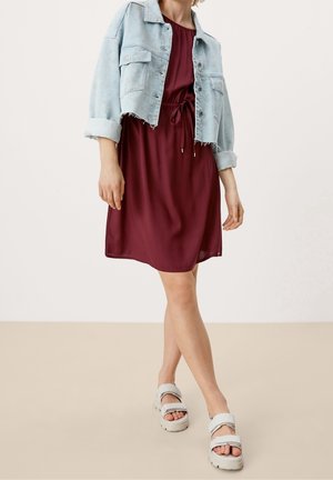 Chaqueta de mezclilla con dobladillo deshilachado, sobre un vestido burdeos con cintura ajustable. La modelo lleva sandalias de plataforma grises. Fondo neutro.