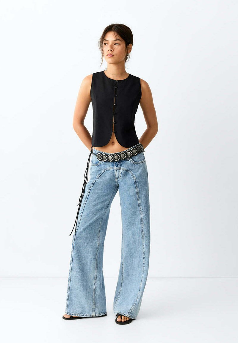 Femme portant un haut noir sans manches boutonné, un jean bleu clair à jambes larges avec des coutures visibles, une ceinture décorative et des sandales noires, debout les mains derrière le dos.