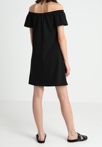 Czarna sukienka typu off-the-shoulder z krótkimi rękawami, sięgająca do kolan, wykonana z miękkiego materiału, z elastycznym dekoltem i luźnym krojem. Noszona z czarnymi sandałami.