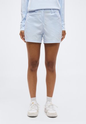 Femme portant un short cintré bleu clair, des baskets blanches, des chaussettes blanches et une chemise à manches longues bleu clair, debout devant un fond uni.