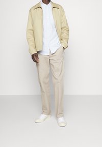 Beżowa kurtka, biała koszula w paski i jasnobeżowe spodnie. Model nosi białe sneakersy. Gładka faktura, nowoczesny design i luźny krój.