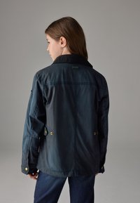 Sort vandtæt jakke med fløjlsagtet krave, trykknaplukning og en afslappet pasform, vist bagfra. Glat tekstur, minimalistisk design.