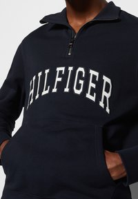 Personne portant un sweat-shirt bleu marine à fermeture éclair quart avec le texte blanc « HILFIGER » et les mains dans la poche avant, sur un fond uni.