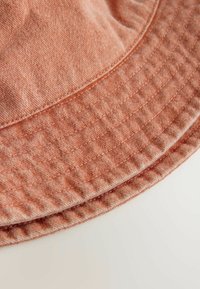Persikofärgad bucket hat tillverkad av strukturerad textil med en sydd band och en bred, rund skärm. Inga synliga märkes- eller logodetaljer.