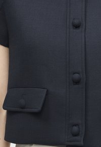 Κοντινό πλάνο σκουρόχρωμου υφασμάτινου ενδύματος με τρία κουμπιά καλυμμένα με ύφασμα και μια μικρή τσέπη με καπάκι και αντίστοιχο κουμπί στην αριστερή πλευρά.