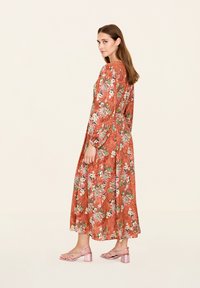 Robe maxi en rouille avec un motif floral, manches longues, poignets élastiques et poches. Associée à des sandales à talons blocs rose clair.