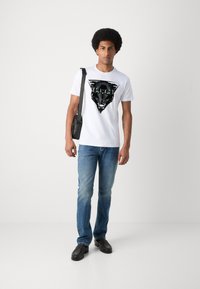Camiseta blanca de algodón con un gráfico de pantera negra y ojos verdes, combinada con jeans de mezclilla azules y zapatos negros. Incluye un bolso negro de hombro.