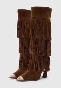 Botas de ante marrón hasta la rodilla con detalles de flecos en capas y una puntera metálica dorada. Diseño de tacón alto, textura suave y forma afilada.