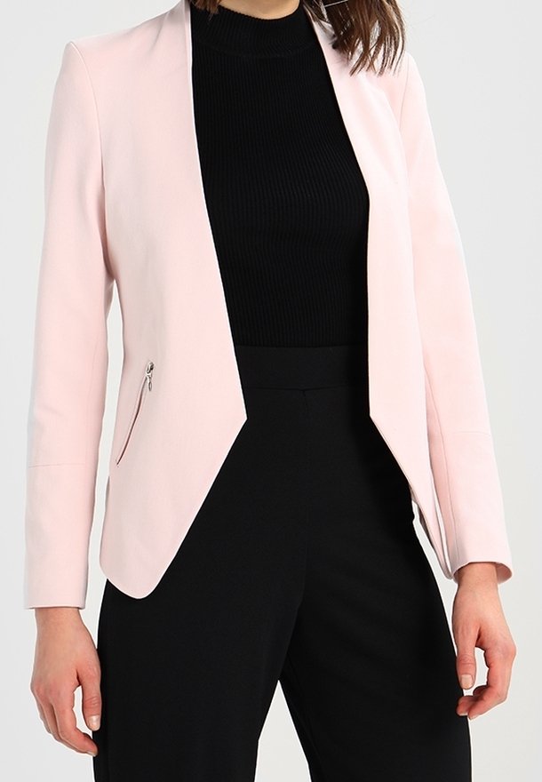 Femme portant un blazer rose clair avec des poches latérales zippées par-dessus un top noir côtelé et un pantalon noir taille haute.