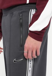 Sive Nike trenirke z zadrgo na stranski žep, teksturirana tkanina in bele akcenti z blagovno znamko "NIKE" na strani.