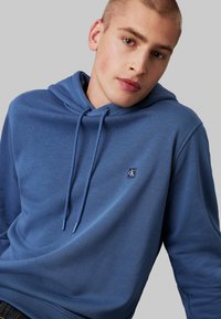 Felpa blu in cotone con cappuccio e coulisse, dotata di un piccolo logo ricamato sul petto e tessuto testurizzato. Finitura liscia e vestibilità rilassata.