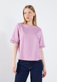 Ljusrosa t-shirt med kort ärm och spetsdetaljer vid ärmsluten, rund hals och avslappnad passform. Slät textur och avslappnad design, kombinerad med mörk denim.