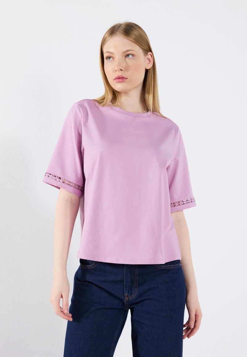 Ljusrosa t-shirt med kort ärm och spetsdetaljer vid ärmsluten, rund hals och avslappnad passform. Slät textur och avslappnad design, kombinerad med mörk denim.
