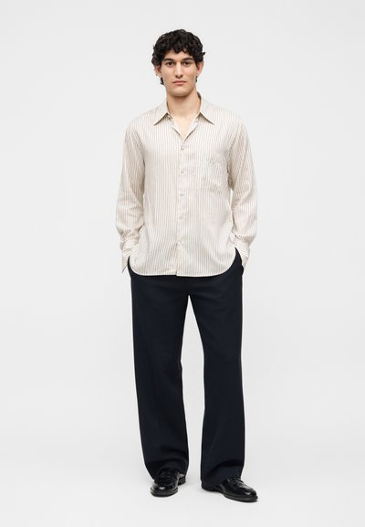Νέος άντρας όρθιος με τα χέρια στις τσέπες, φοράει μπεζ ριγέ μακρυμάνικο πουκάμισο, μαύρο παντελόνι με φαρδιά πόδια και μαύρα παπούτσια σε λευκό φόντο.
