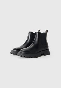 Une paire de bottes Chelsea en cuir noir avec des semelles épaisses à motif relief et des panneaux latéraux élastiques sur un fond blanc uni.
