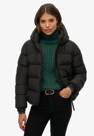 Zwarte pufferjas met een capuchon, ritssluiting en elastische manchetten, gedragen over een groene gebreide coltrui en zwarte jeans met een riem.