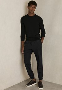 Cămașă lungă neagră cu mâneci lungi, asociată cu pantaloni jogger negri tapered. Pantalonii au manșete elastice. Purtați cu pantofi slip-on de culoare maro închis.