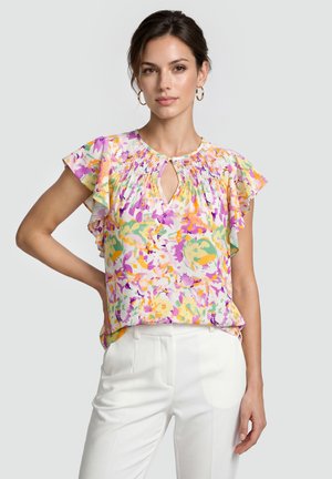 Kvinde iført en farverig blomstret bluse med flagrende ærmer og hvide bukser, stående med den ene hånd på hoften mod en ensfarvet baggrund.