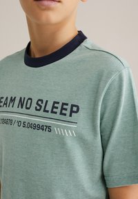 Zelené tričko s krátkym rukávom a námorníckym golierom, s nápisom "DREAM NO SLEEP" a grafickými detailmi v tmavomodrej a bielej farbe.
